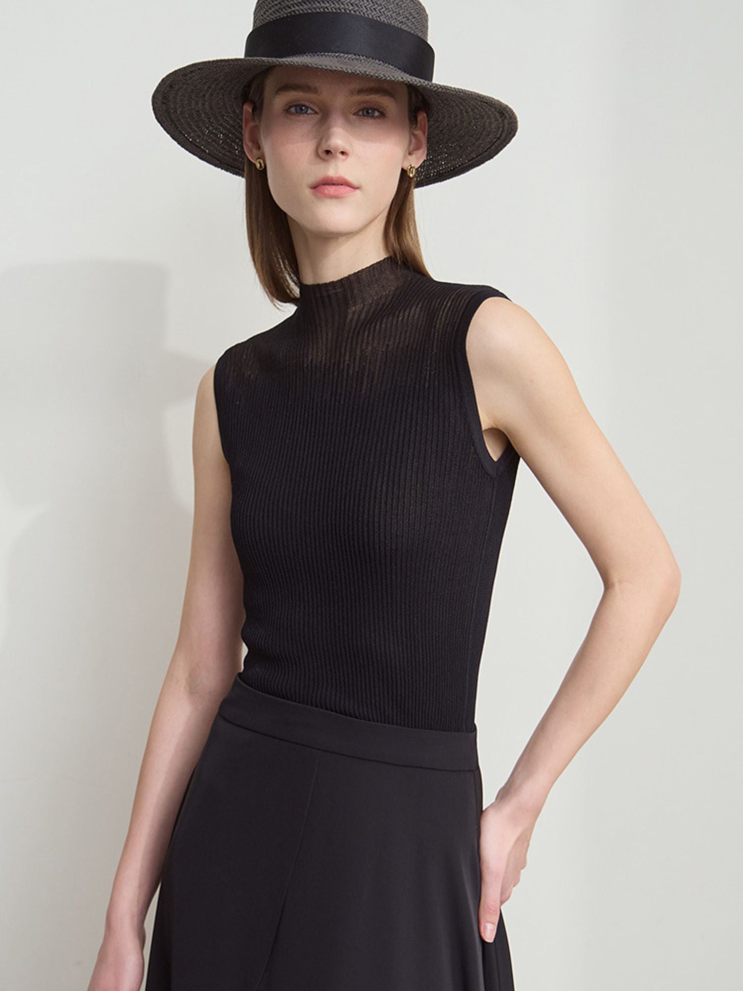 Frau trägt ärmelloses schwarzes Stricktop mit Stehkragen und weitem Rock, kombiniert mit schwarzem Hut