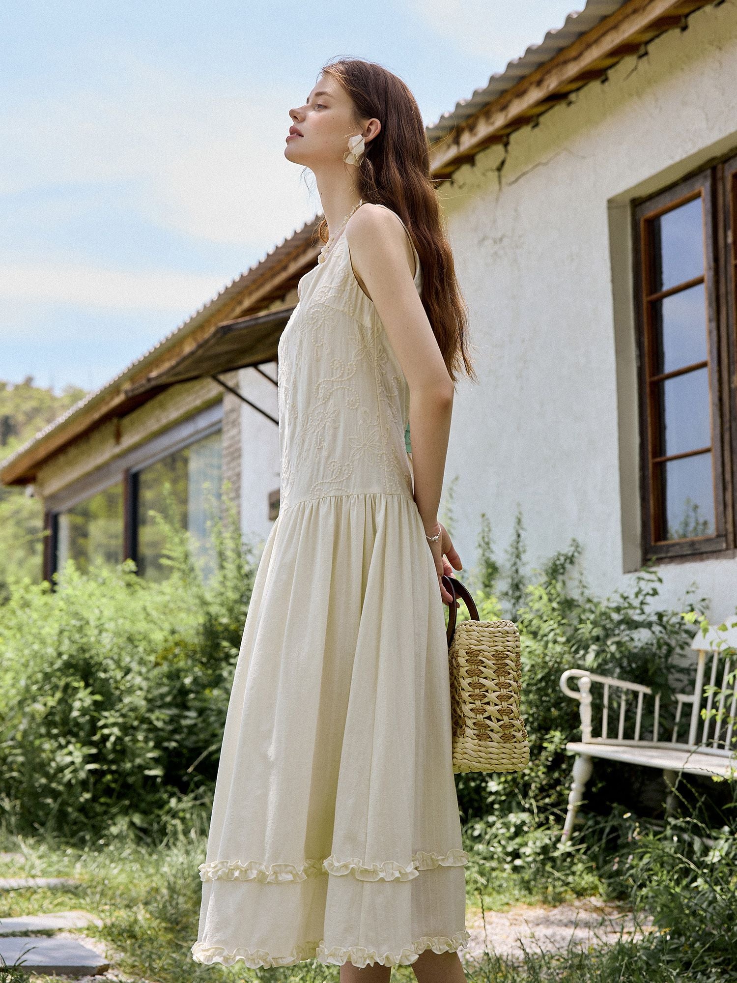 Modell trägt ein ärmelloses cremefarbenes Midikleid mit geraffter Taille und Spitzenbesatz, dazu eine Korbtasche im Garten