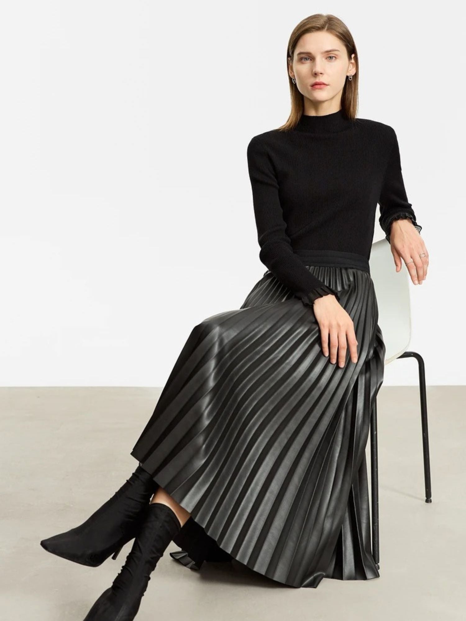 Model seated in black pleated faux leather midi skirt with ribbed black sweater and boots