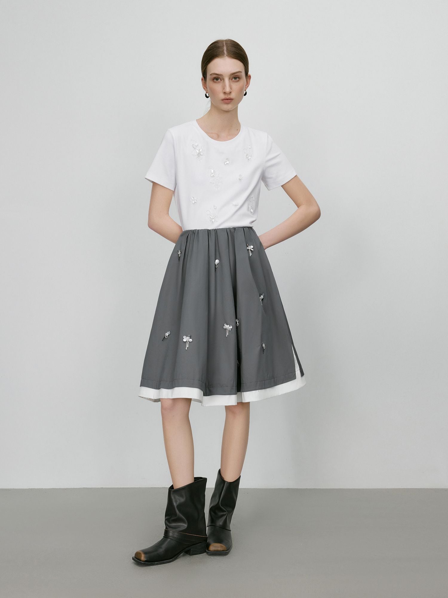 Frau trägt weißen T-Shirt mit grauem Midirock mit Blumenstickerei und schwarzen Stiefeletten
