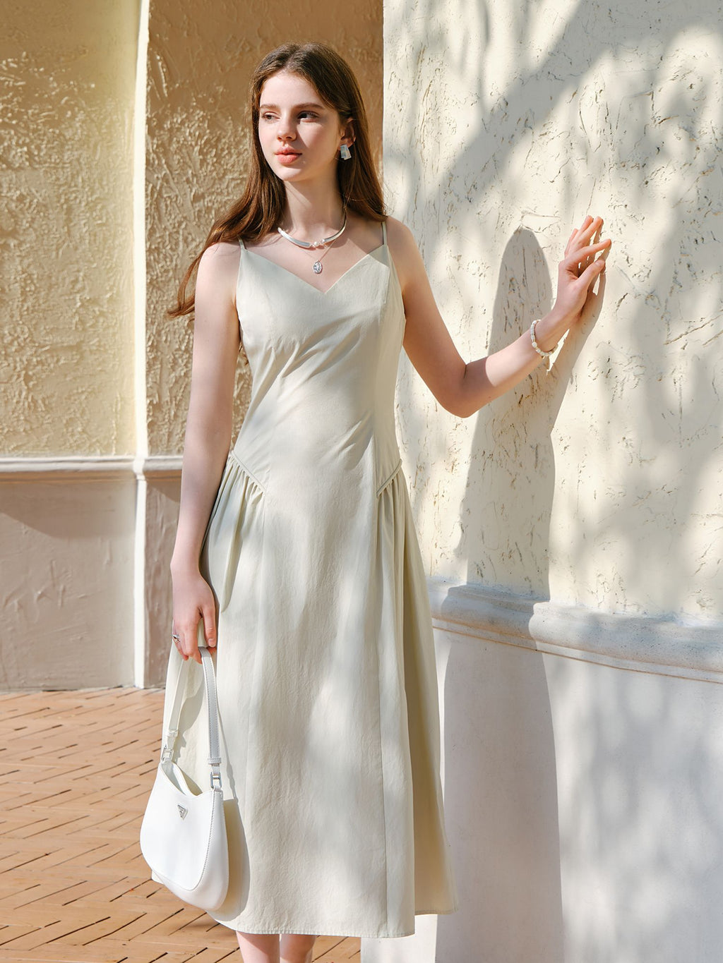 Modell im weißen Trägerkleid mit V-Ausschnitt und Baguette, stehend im Sonnenlicht an der Wand