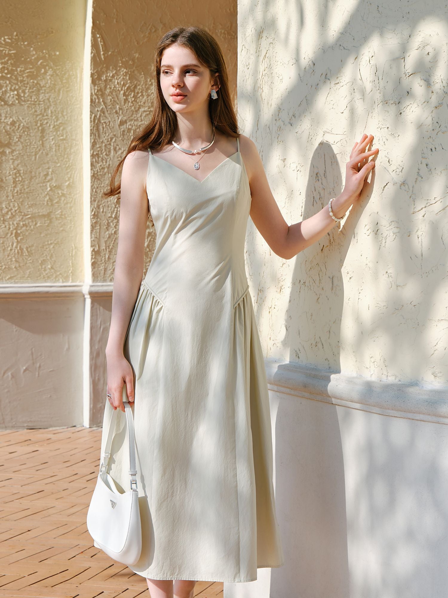 Modell im weißen Trägerkleid mit V-Ausschnitt und Baguette, stehend im Sonnenlicht an der Wand