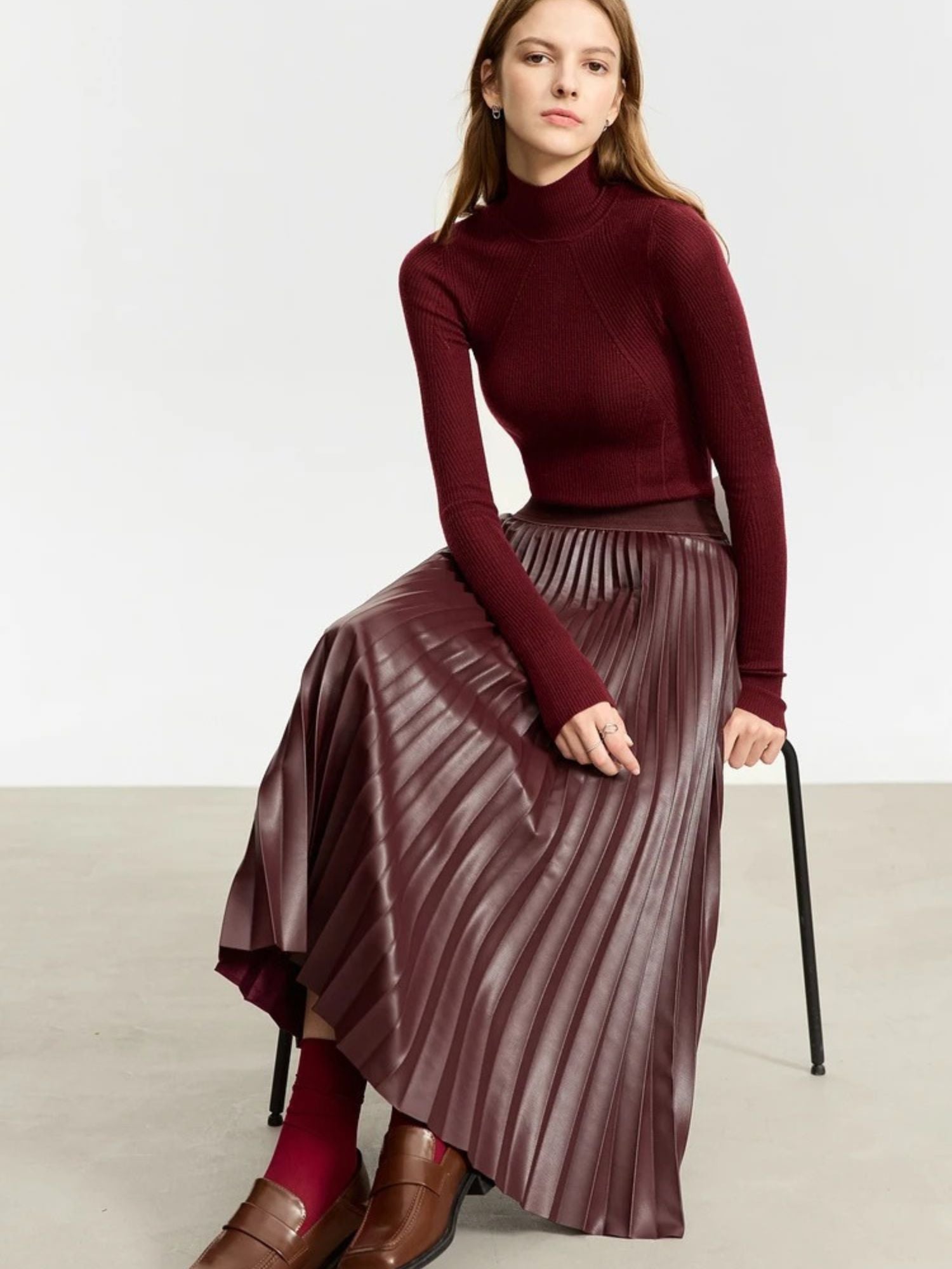 Model seated in burgundy pleated faux leather skirt with long-sleeve turtleneck
